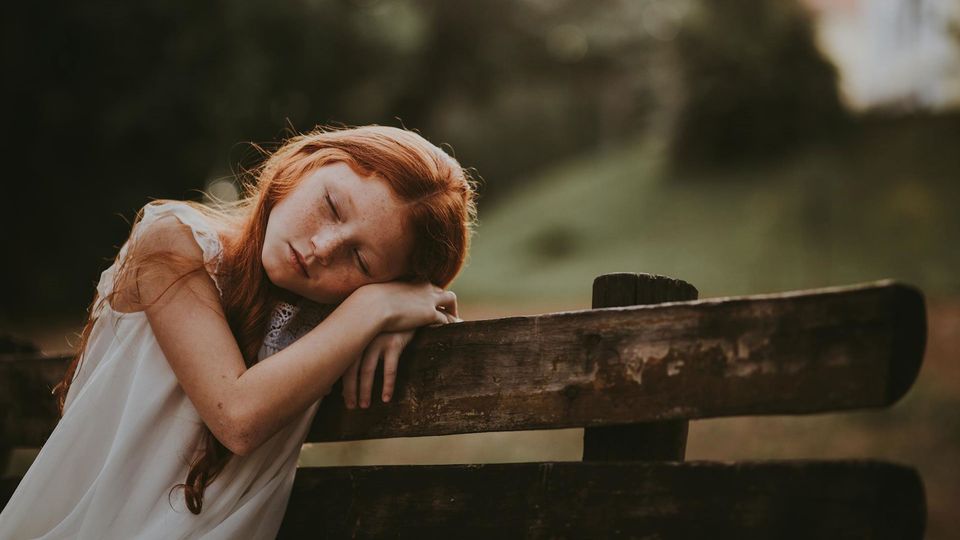 A child rests her head on her hands while sat on a bench.